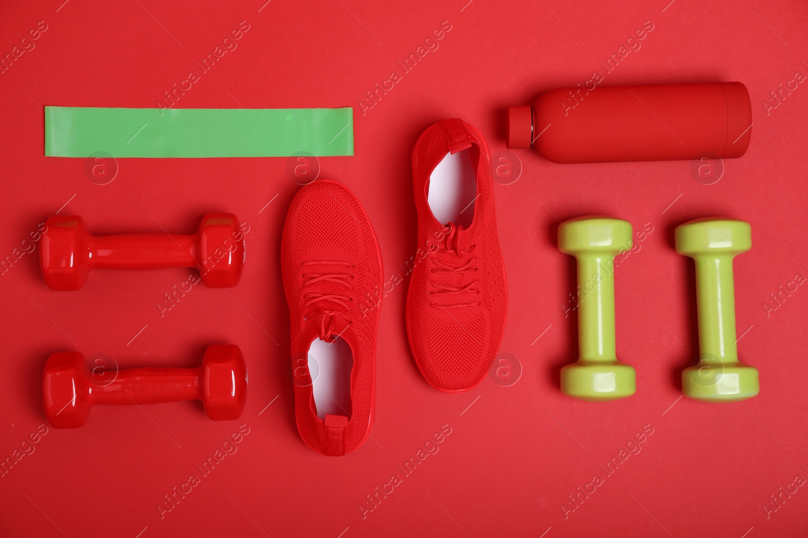 Photo of Elastic band, water bottle, dumbbells and sneakers on red background, flat lay. Sports equipment