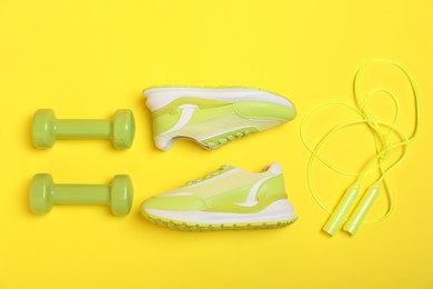 Photo of Dumbbells, skipping rope and sneakers on yellow background, flat lay. Sports equipment