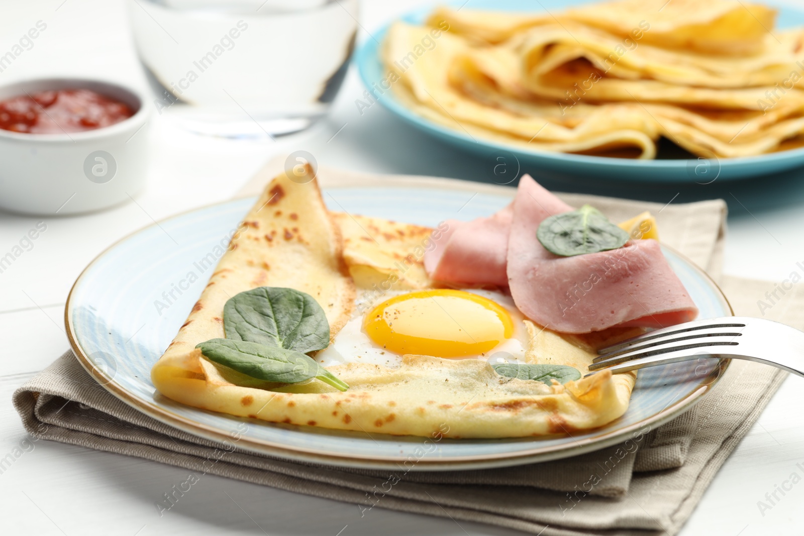 Photo of Delicious crepes with egg, ham, basil and sauce on white wooden table, closeup