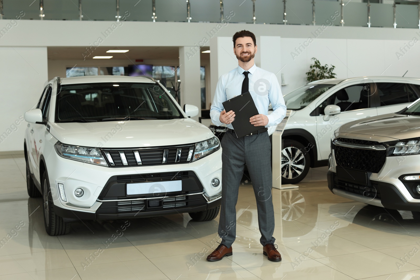 Photo of Happy salesman with clipboard near new cars in salon