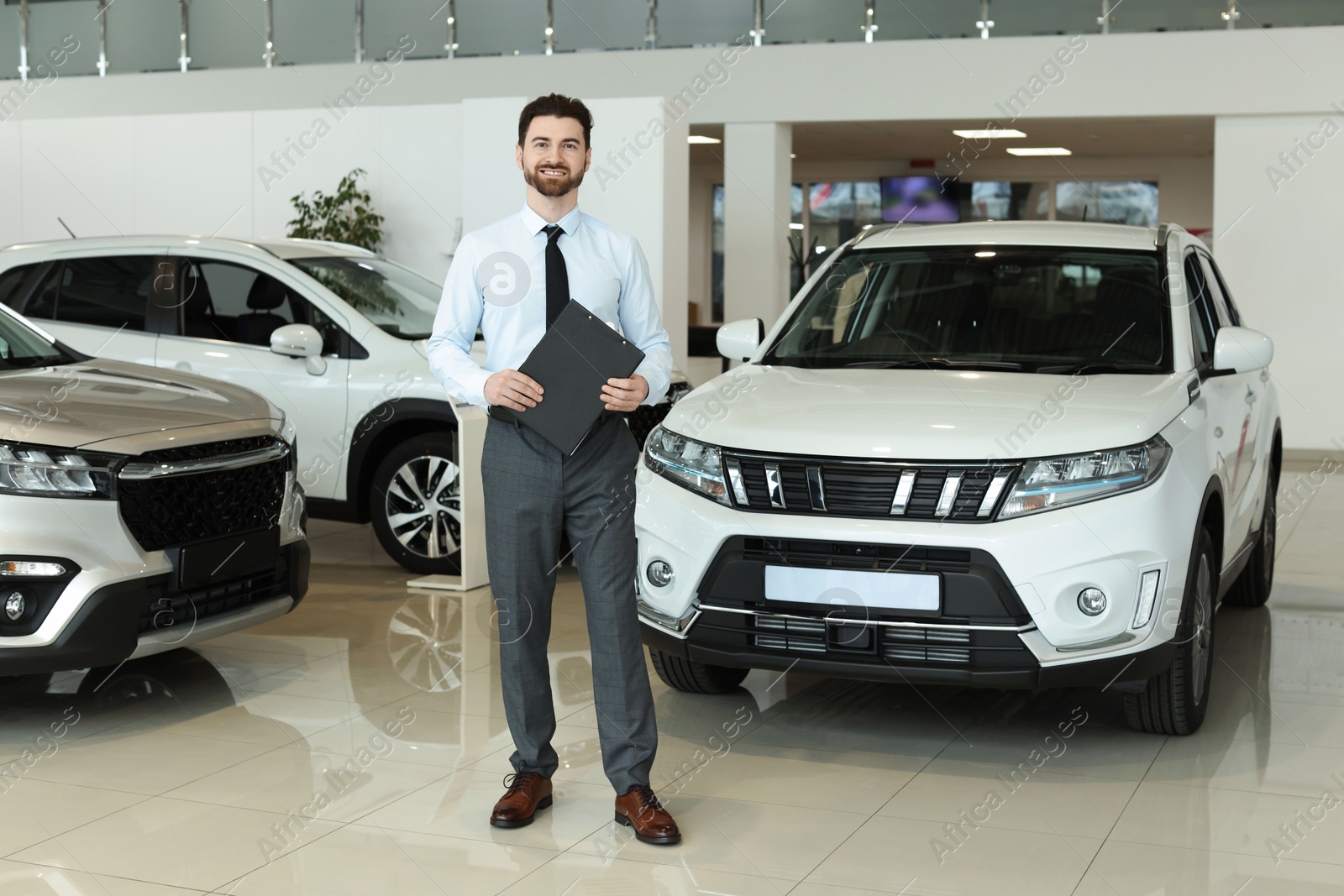 Photo of Happy salesman with clipboard near new cars in salon