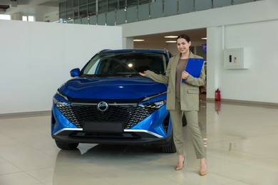 Happy saleswoman with clipboard near new blue car in salon