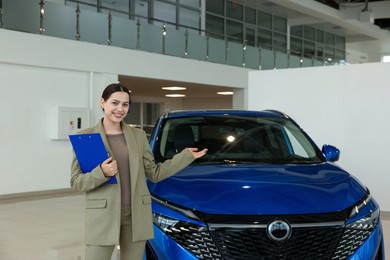 Photo of Happy saleswoman with clipboard near new blue car in salon