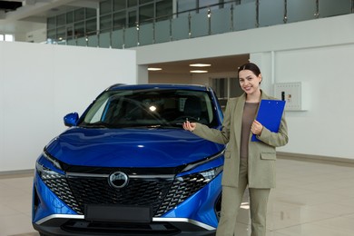 Happy saleswoman with clipboard near new blue car in salon