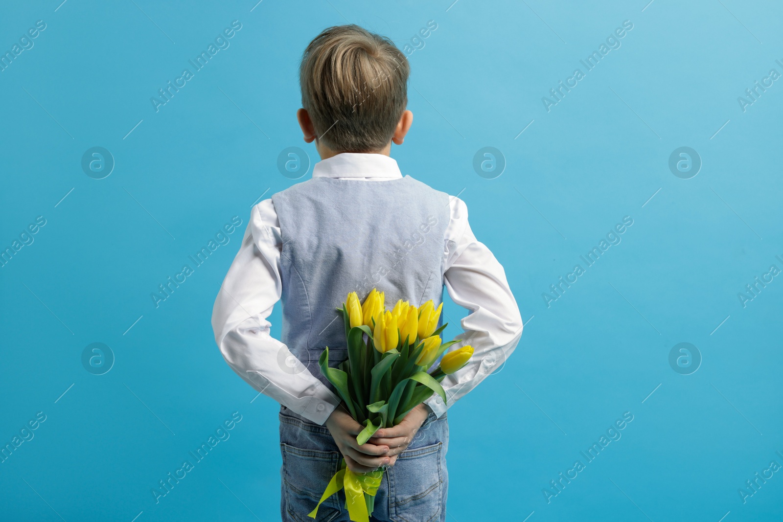 Photo of Little boy with bouquet of tulips behind his back on light blue background