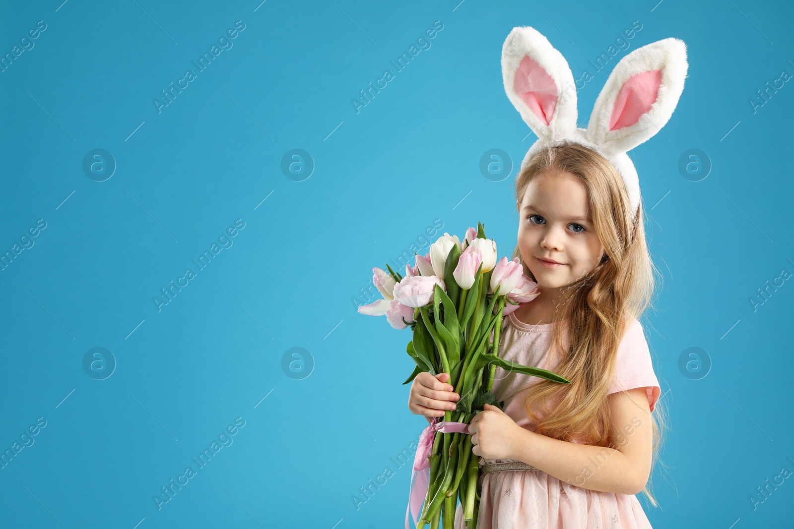 Photo of Cute little girl with bunny ears and spring flowers on light blue background, space for text. Easter celebration