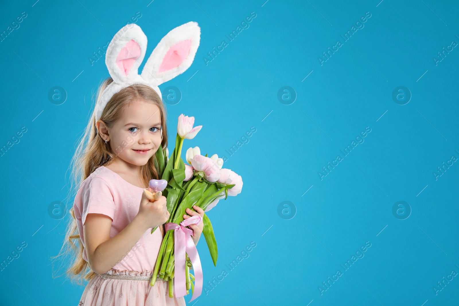 Photo of Cute little girl with bunny ears and spring flowers on light blue background, space for text. Easter celebration