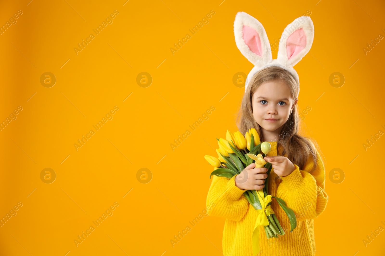 Photo of Cute little girl with bunny ears and tulips on orange background, space for text. Easter celebration