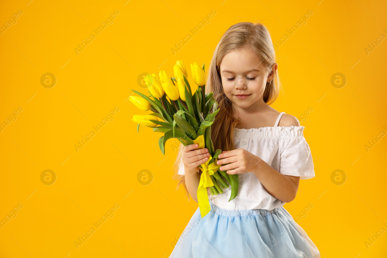 Photo of Cute little girl with beautiful yellow tulips on orange background, space for text. Spring season