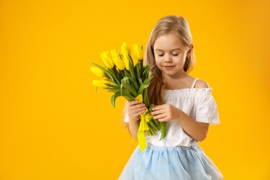 Photo of Cute little girl with beautiful yellow tulips on orange background, space for text. Spring season