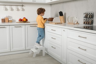 Photo of Little boy with cookies standing on step stool near countertop in kitchen