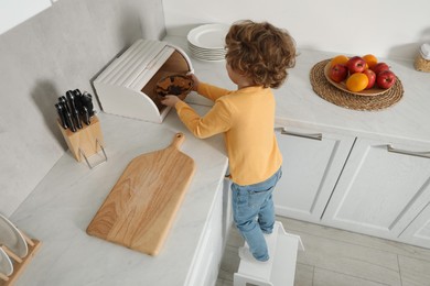 Photo of Little boy standing on step stool and reaching for cookies in kitchen