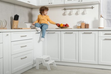 Photo of Little boy taking apple on countertop and step stool in kitchen