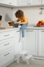 Photo of Little boy taking cookie on countertop and step stool in kitchen