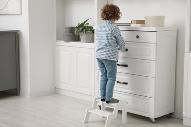 Photo of Little boy standing on step stool near chest of drawers at home, back view