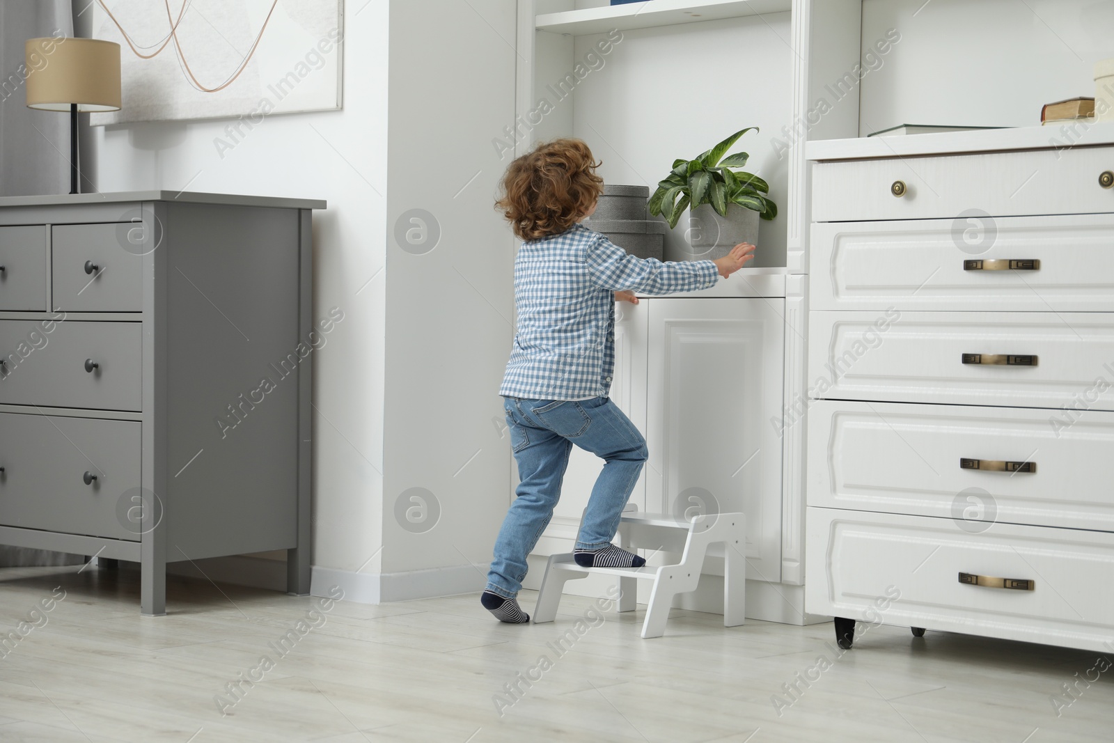 Photo of Little boy standing on step stool near shelving unit at home