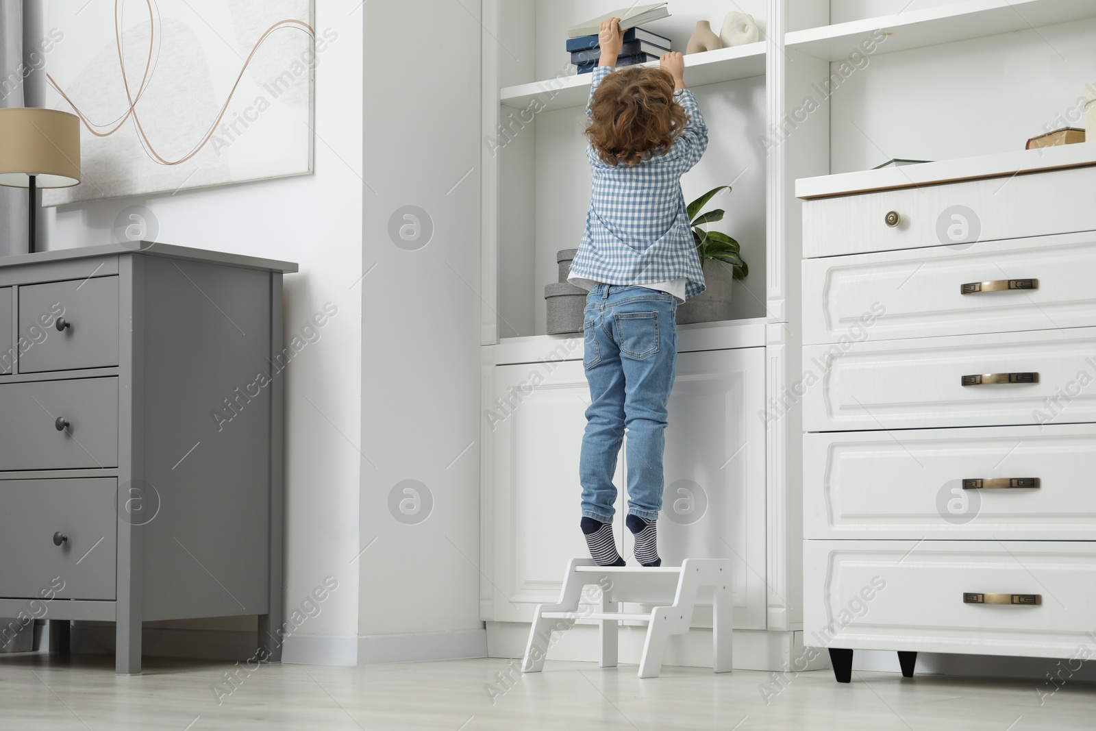 Photo of Little boy standing on step stool and reaching for book at home, back view