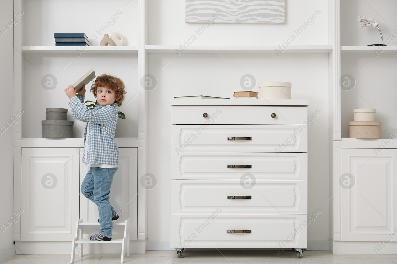 Photo of Little boy with book standing on step stool near shelving unit at home