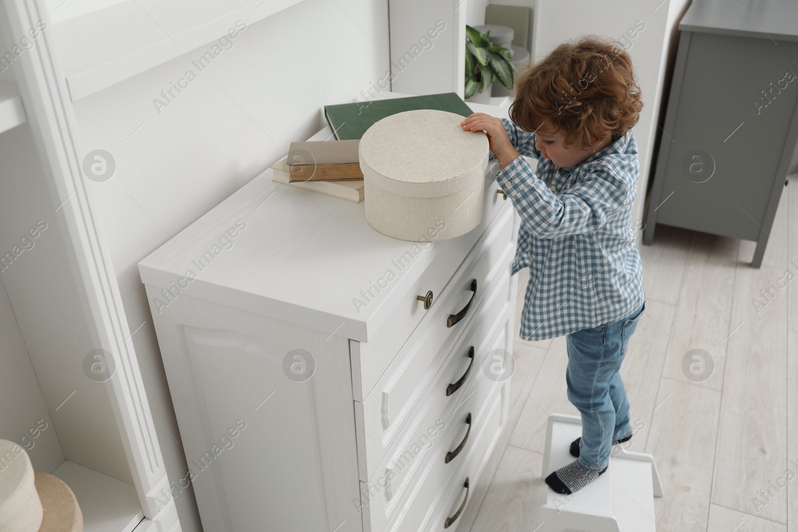 Photo of Little boy standing on step stool and reaching for box at home