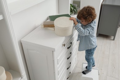 Photo of Little boy standing on step stool and reaching for box at home