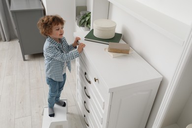 Photo of Little boy standing on step stool near chest of drawers at home