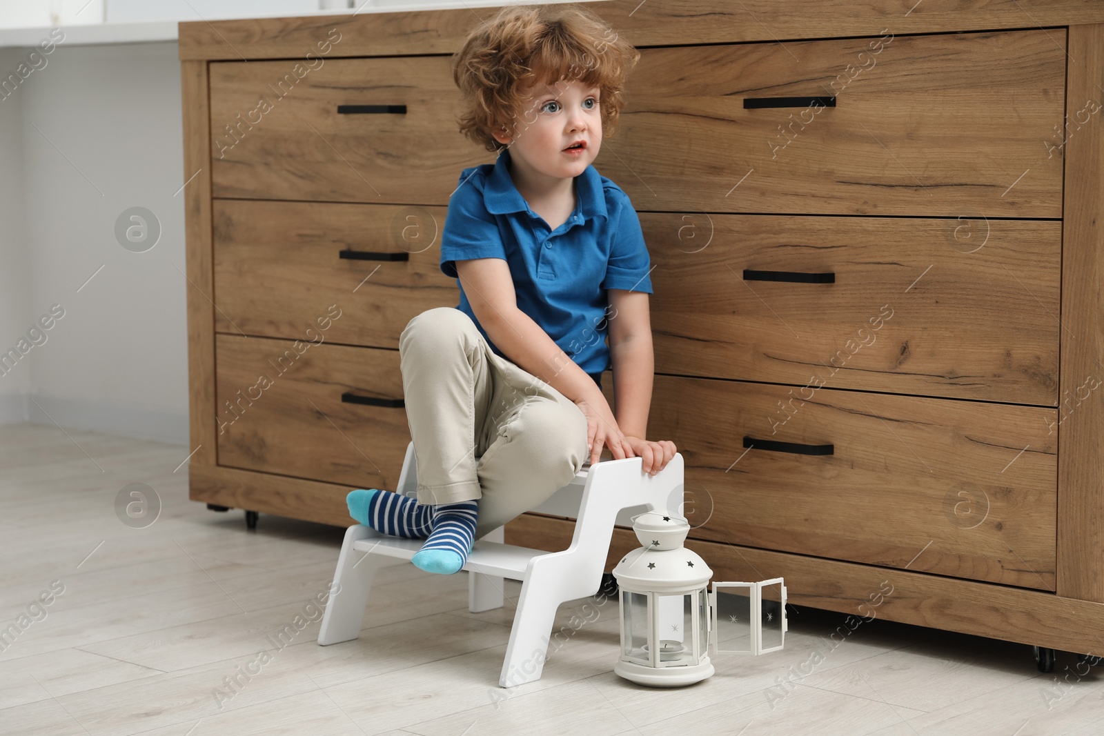 Photo of Little boy sitting on step stool near chest of drawers at home