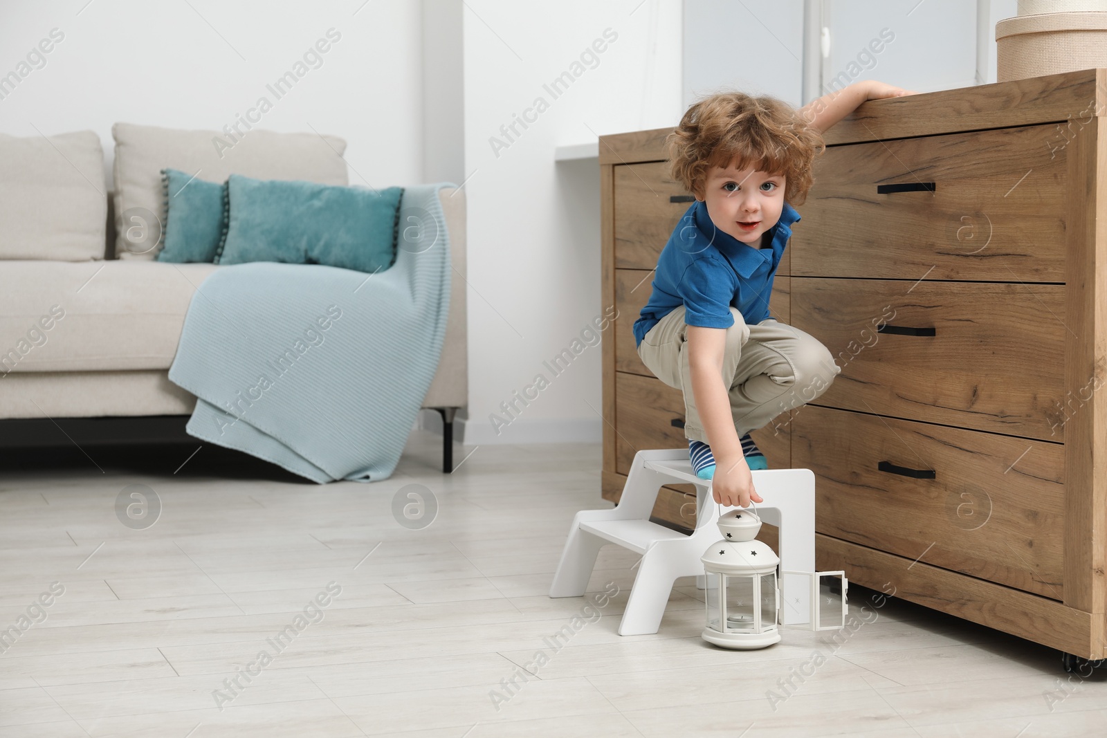 Photo of Little boy with lantern standing on step stool near chest of drawers at home