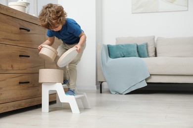 Photo of Little boy with box standing on step stool near chest of drawers at home