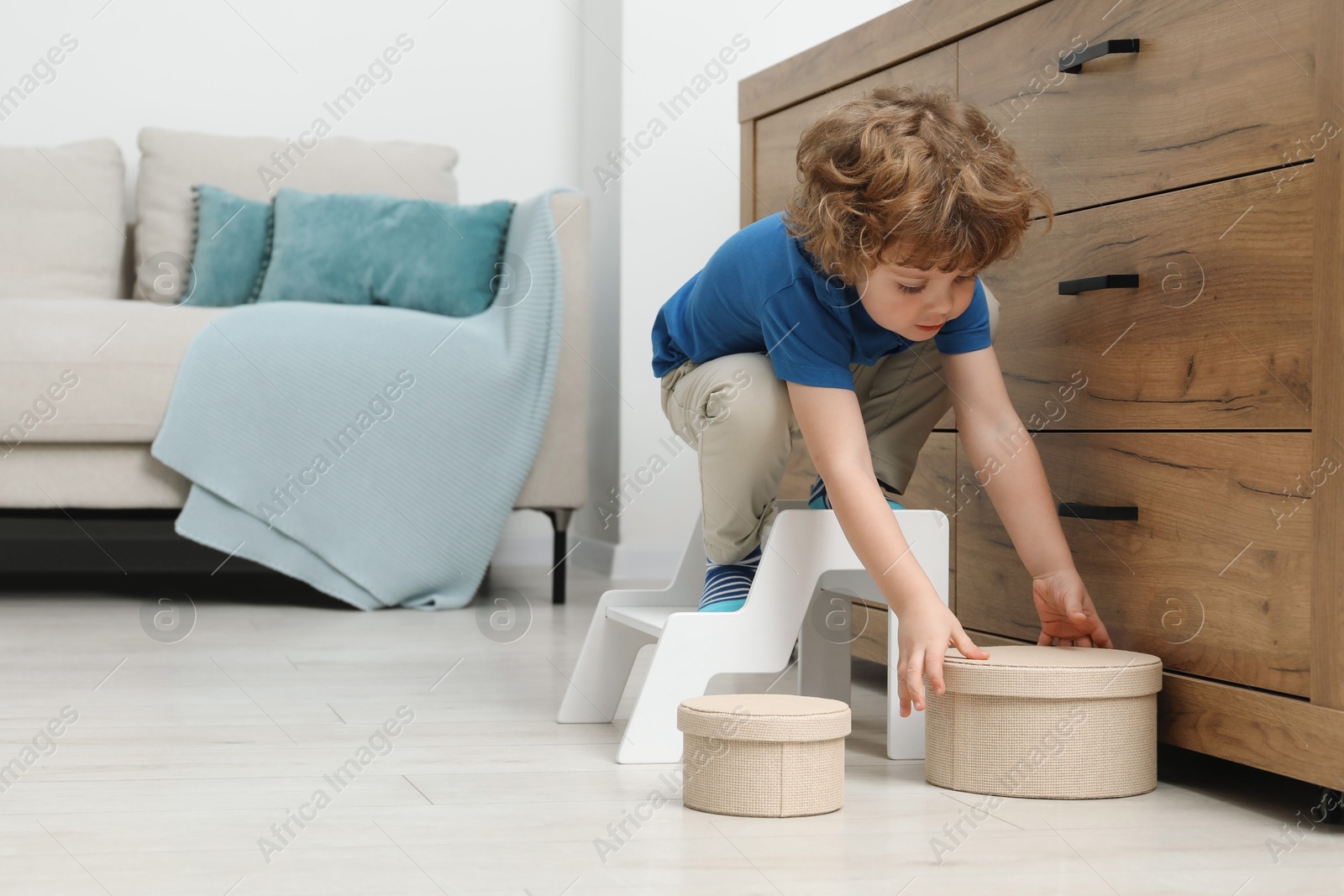 Photo of Little boy reaching for box while standing on step stool near chest of drawers at home