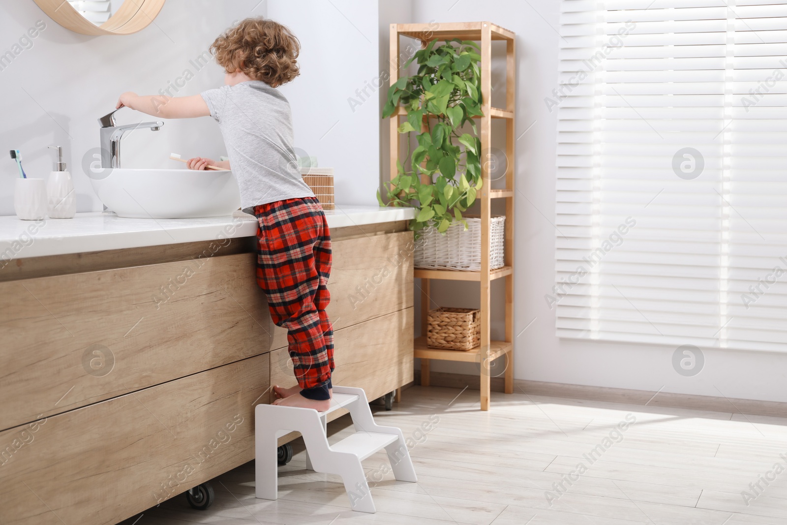 Photo of Little boy with toothbrush standing on step stool near bathroom vanity indoors