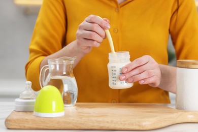 Photo of Mother making baby formula in feeding bottle at table indoors, closeup