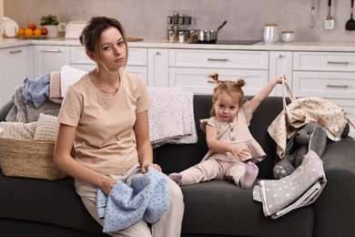 Photo of Tired housewife with her little daughter among messy laundry on sofa at home