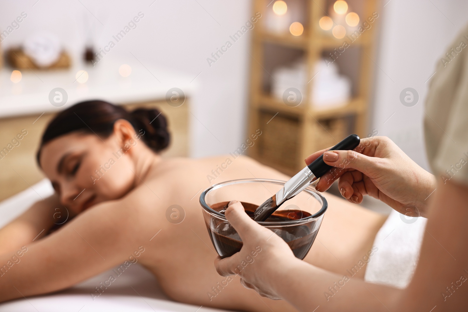 Photo of Spa worker with cosmetic product for chocolate body wrap near woman in salon, closeup. Selective focus