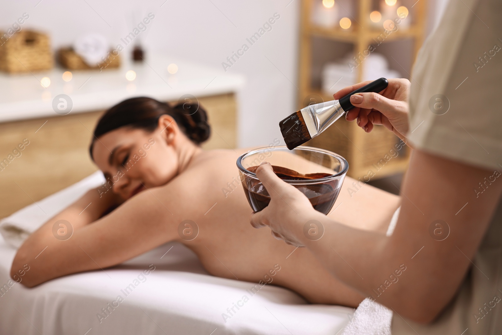 Photo of Spa worker with cosmetic product for chocolate body wrap near woman in salon, closeup. Selective focus