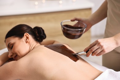 Photo of Chocolate body wrap. Spa worker applying mask onto woman's back in salon, closeup