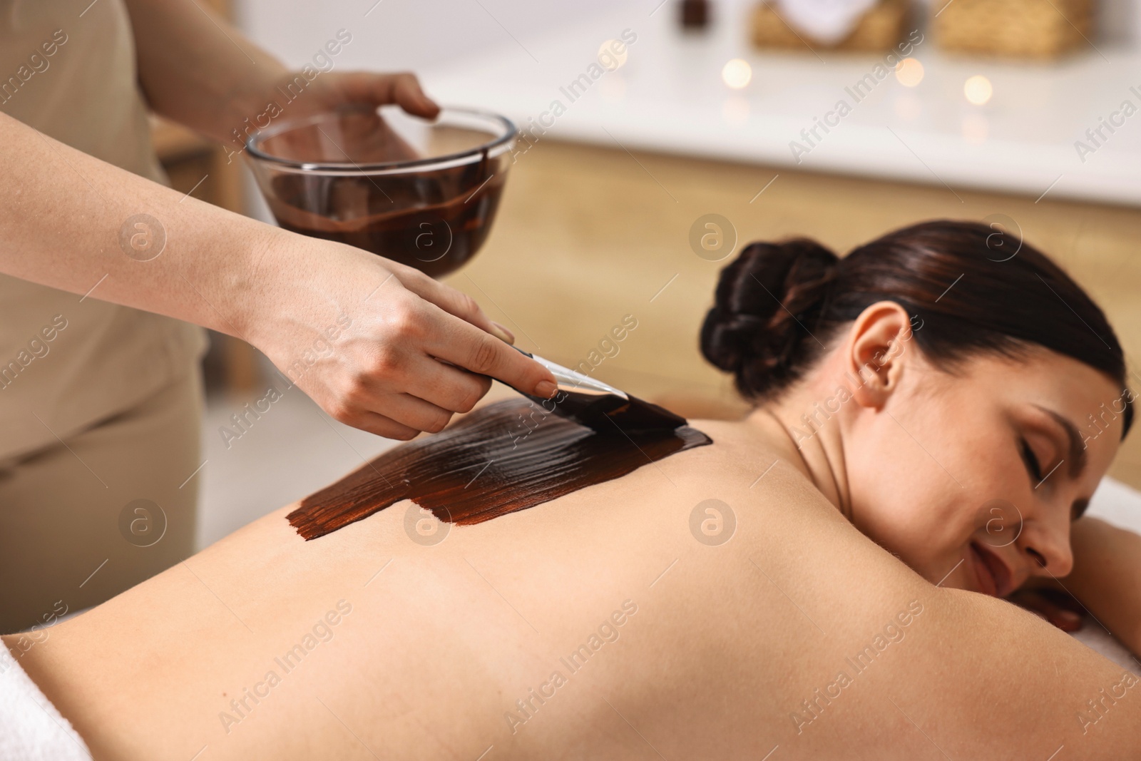 Photo of Chocolate body wrap. Spa worker applying mask onto woman's back in salon, closeup