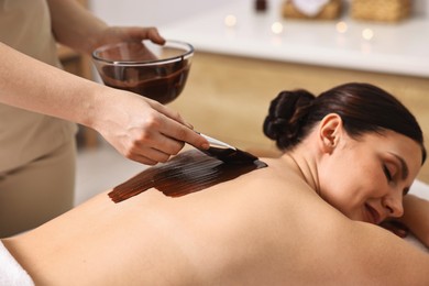 Photo of Chocolate body wrap. Spa worker applying mask onto woman's back in salon, closeup