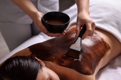 Photo of Chocolate body wrap. Spa worker applying mask onto woman's back in salon, closeup