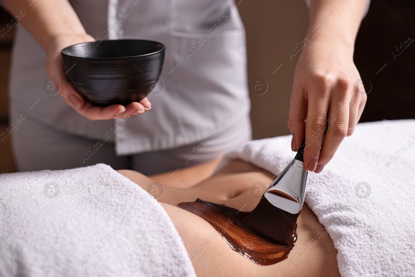 Photo of Chocolate body wrap. Spa worker applying mask onto woman's belly in salon, closeup