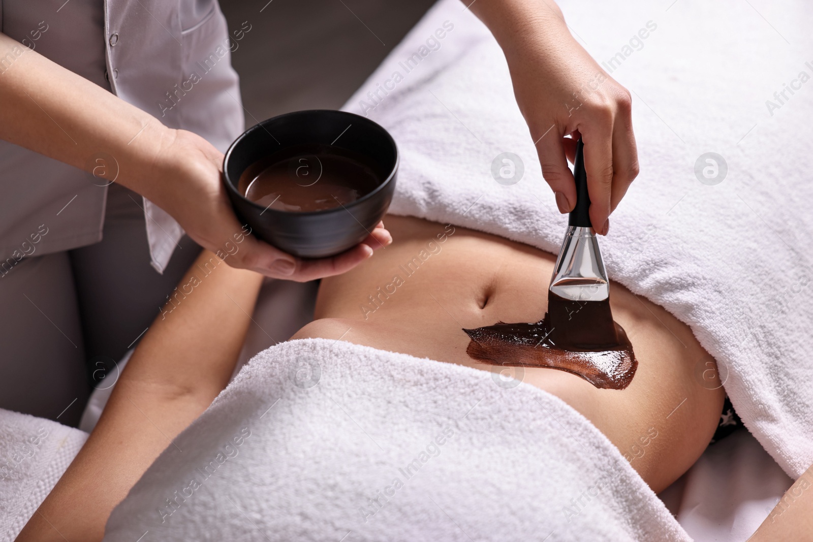 Photo of Chocolate body wrap. Spa worker applying mask onto woman's belly in salon, closeup