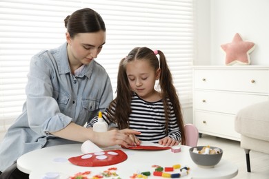 Photo of Woman and little girl making paper fly agaric at white table indoors. Child handmade craft