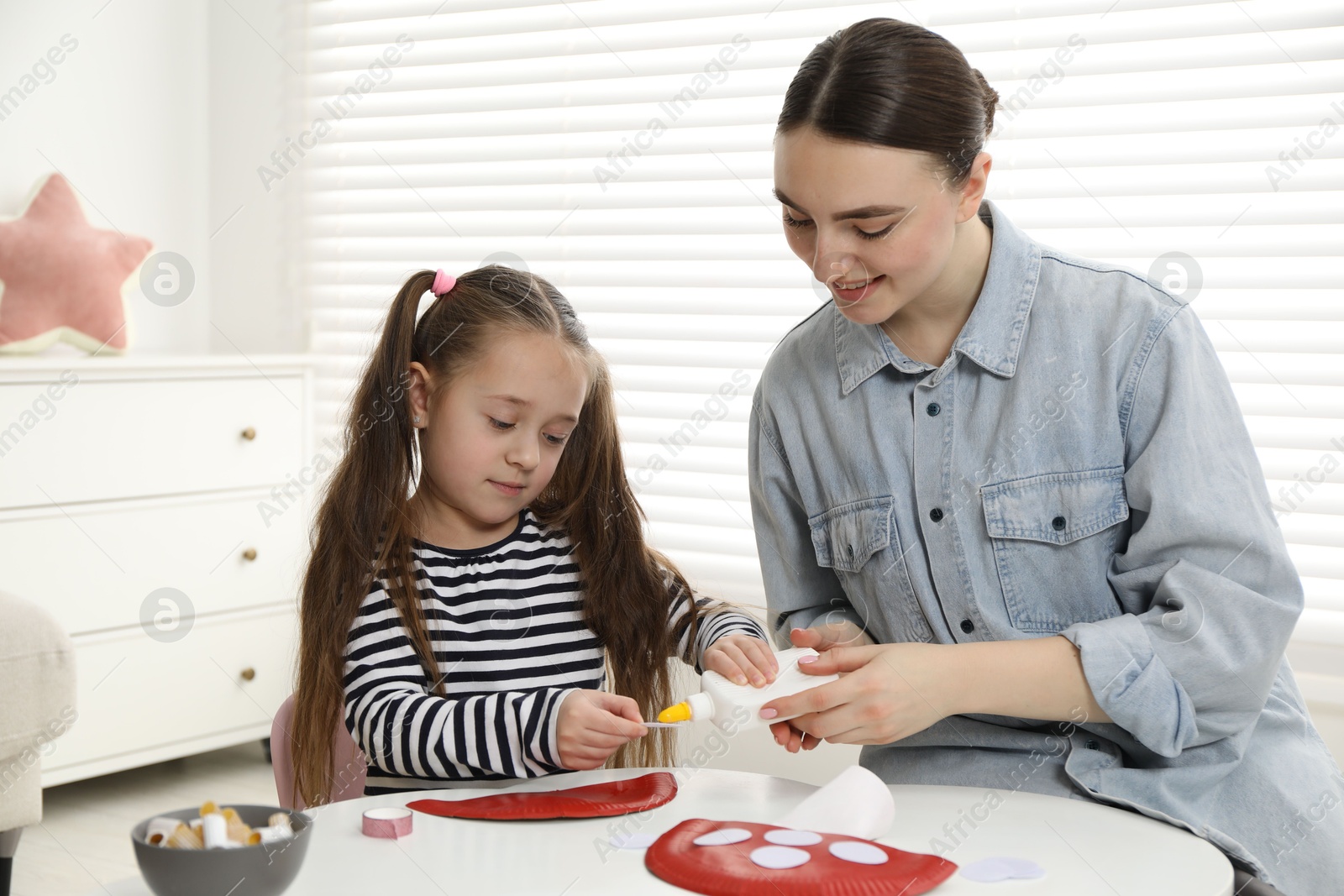 Photo of Woman and little girl making paper fly agaric at white table indoors. Child handmade craft