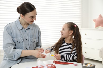 Photo of Woman and little girl making paper fly agaric at white table indoors. Child handmade craft