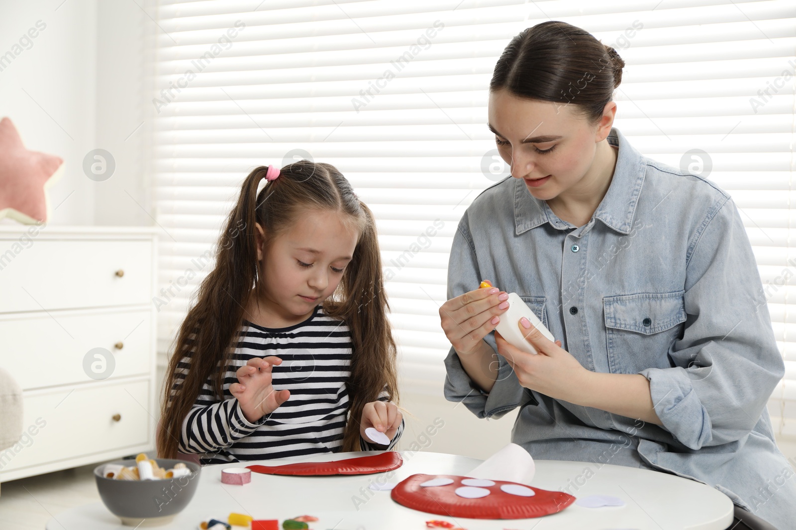 Photo of Woman and little girl making paper fly agaric at white table indoors. Child handmade craft