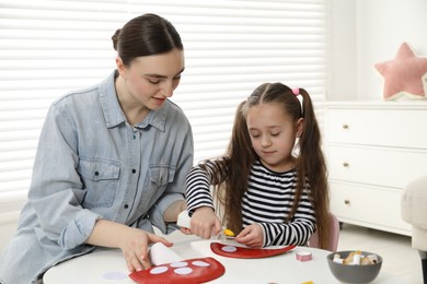 Photo of Woman and little girl making paper fly agarics at white table indoors. Child handmade craft