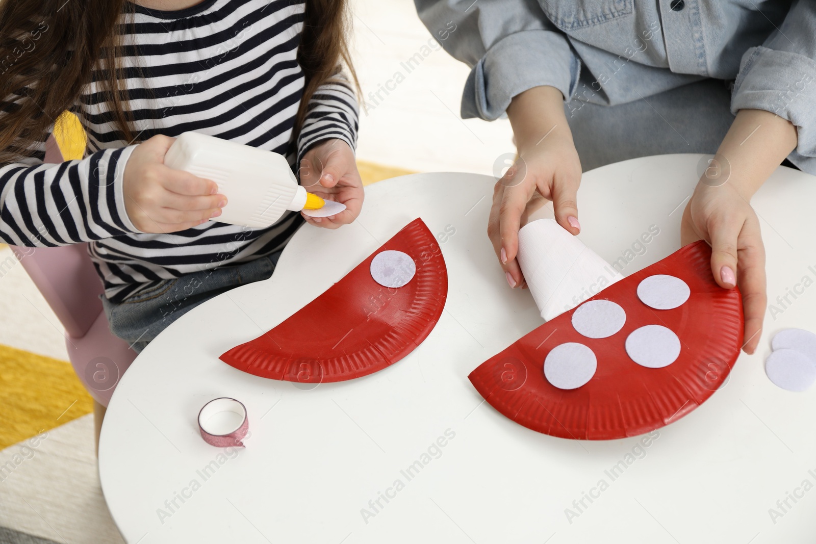 Photo of Woman and little girl making paper fly agarics at white table indoors, closeup. Child handmade craft