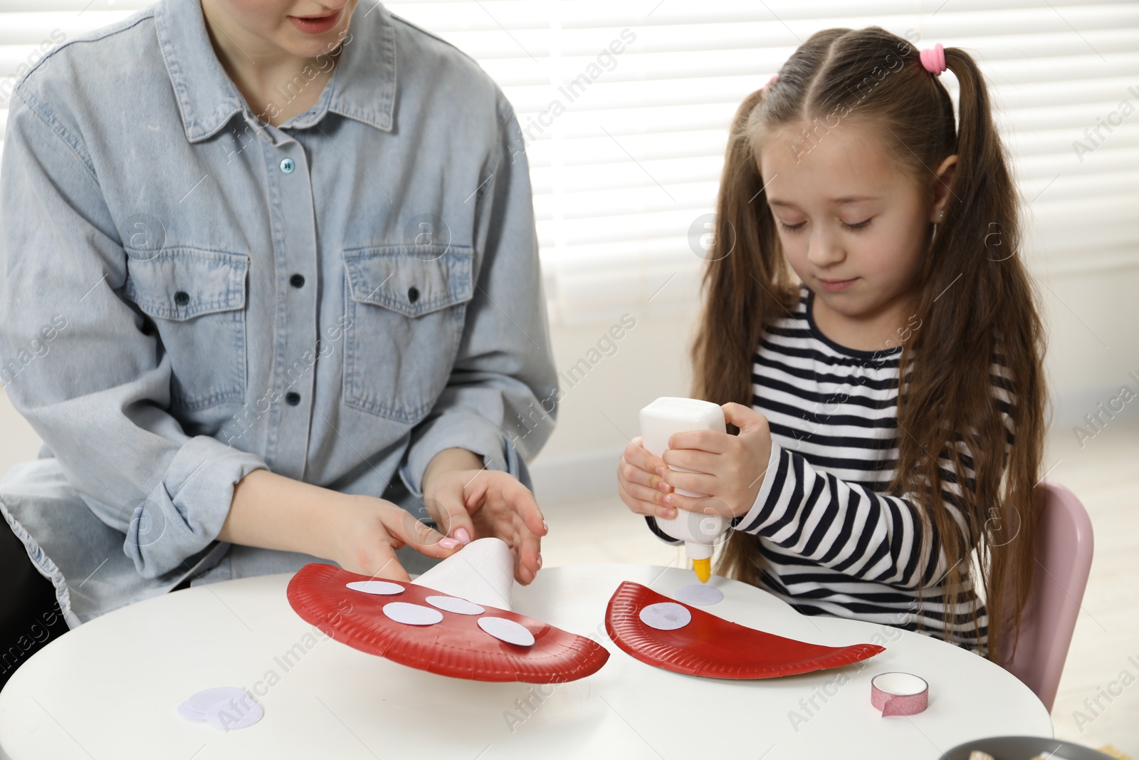 Photo of Woman and little girl making paper fly agarics at white table indoors, closeup. Child handmade craft