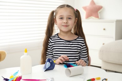 Photo of Little girl making paper bird at white table indoors. Child handmade craft