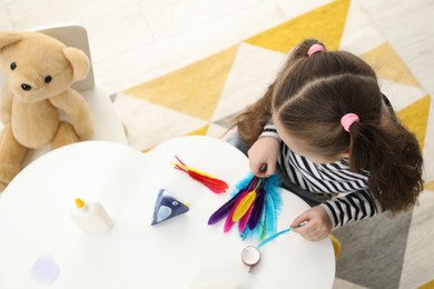 Photo of Little girl making paper bird at white table indoors, top view. Child handmade craft