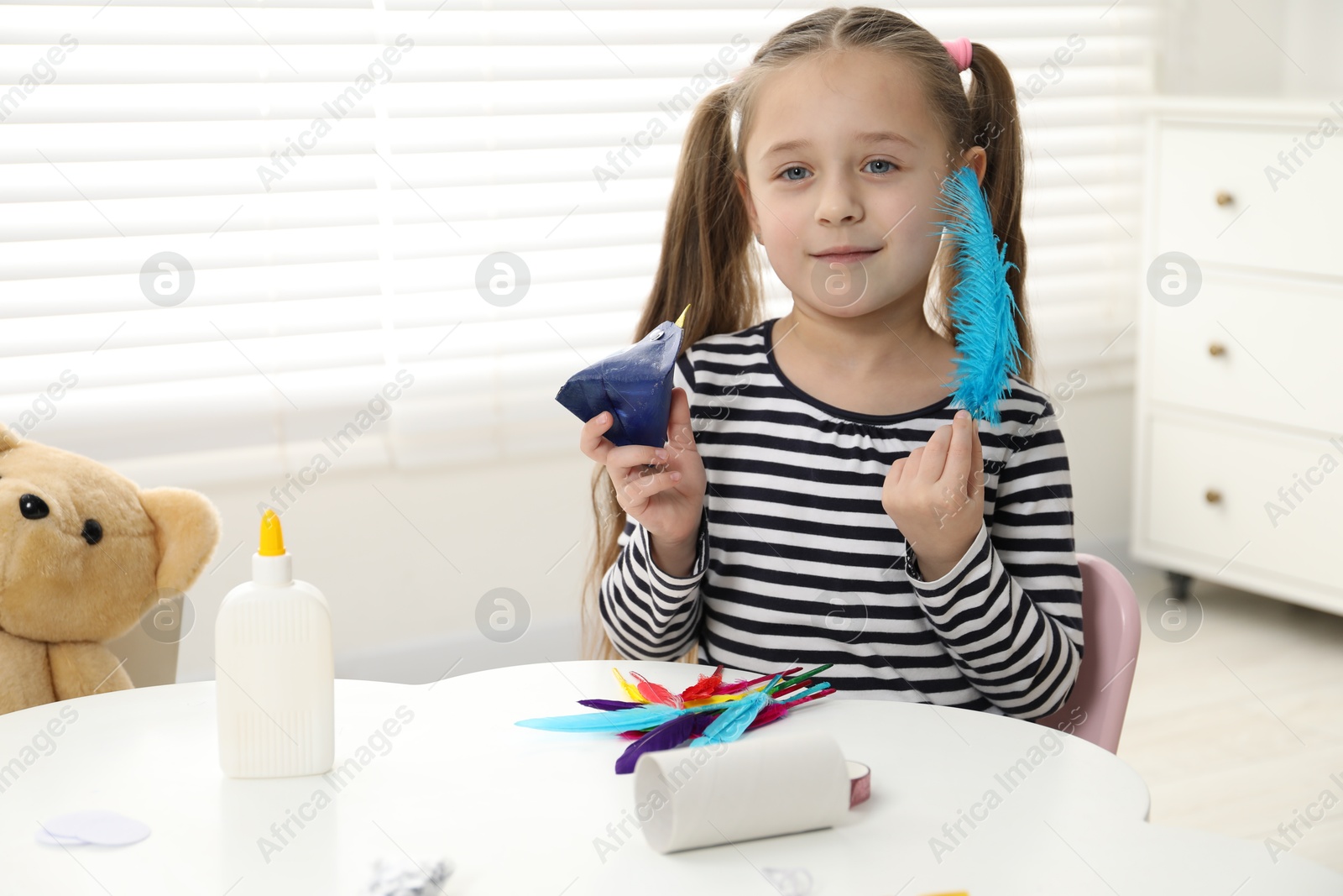 Photo of Little girl with paper bird and light blue feather indoors. Child handmade craft